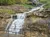 A small waterfall flows under a metal bridge in a forest, with a large spherical sculpture nearby.