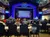 The image shows a theater or performance venue with an audience seated at tables facing a stage, which is adorned with an elaborate frame and a projected screen.