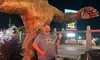 A man is posing with a green golf club in front of a large Bigfoot statue at night.
