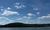 The image shows a scenic view of a lake with distant tree-covered hills under a bright blue sky, observed from a boat deck with some people in view.
