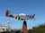 A WWII-era fighter plane replica is mounted on a brick pedestal against a clear blue sky, with a parking lot and American flags in the background.