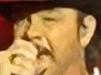 A man and woman are performing on stage in front of a vibrant red and white abstract background, with a drummer and guitarist in the scene.