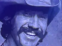 Three performers on stage, with one playing a guitar, are standing in front of a large screen displaying an image of a smiling man wearing a cowboy hat.