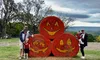A family poses in front of large hay bales painted like jack-o'-lanterns, set against a backdrop of trees and cloudy skies.