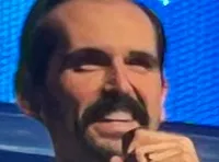 A group of performers dressed in white and gold outfits is singing on a brightly lit stage with a festive backdrop.