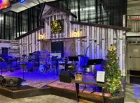 The image shows a rustic music stage set up with microphones, instruments, and a festive Christmas tree beside a wooden barn-style wall adorned with lights and a wreath.