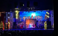 A Christmas theatrical performance is taking place on stage, featuring a nativity scene with several actors, choir members dressed as angels, and festive decorations including Christmas trees.