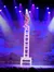 A performer balances on a tall stack of chairs on stage, illuminated by colorful spotlights against a backdrop with cloud designs.