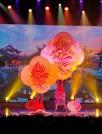 Performers on stage are balancing large, colorful fans on their feet against a backdrop of traditional architecture and vibrant lighting.