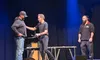 Three men are on a stage, with one man appearing to hand a metal object to another, while the third observes.