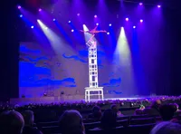 An acrobat is performing a balancing act on a stacked chair tower on a stage under colorful spotlights, viewed by an audience.