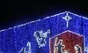 The image features a Christmas nativity scene outlined in colorful lights on a structure, with figures such as Mary, Joseph, and baby Jesus, set beneath a starry background.
