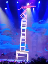 A performer balances acrobatically on a stack of chairs on stage under colorful lights.