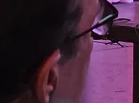 Three performers in colorful costumes are on stage in a rustic, festive setting, with an audience watching.