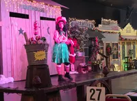 A performer in a whimsical costume stands on a stage decorated with a festive old-time village theme.