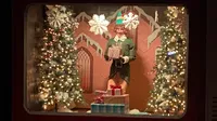 A festive window display featuring a person dressed as an elf holding a gift, surrounded by Christmas trees, lights, and large snowflakes.