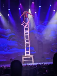 A performer balances on a chair stack during a stage show with colorful, dramatic lighting.