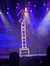 A performer balances on a chair stack during a stage show with colorful, dramatic lighting.