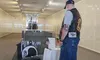 A man wearing a vest and cap stands in front of a POW-MIA tribute display in a dimly lit room.