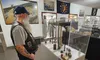 A man with a long beard and cap is observing a museum exhibit featuring military artifacts, including a machine gun and ammunition.