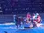 A festive scene with a horse-drawn sleigh featuring people in colorful costumes, including a person dressed as Santa Claus, is performing in front of an audience in an indoor arena.