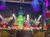 A festive group of people in elf costumes perform energetically on stage, with a large Christmas tree as the backdrop and colorful lighting illuminating the scene.