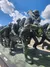 The image shows a sculpture of soldiers in motion, carrying equipment, set against a backdrop of a bright blue sky with scattered clouds.