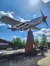A person stands next to a large airplane monument mounted on a brick pedestal in an outdoor memorial setting.