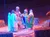 The image shows a theatrical performance depicting a nativity scene with actors in colorful costumes.