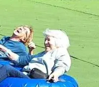 Two people are joyfully sliding down a grassy hill on a blue inflatable tube.