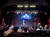 An audience sits in front of a stage featuring a large screen displaying a colorful graphic, surrounded by stage lighting and equipment.