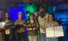 A group of five people pose happily in a room with colorful lighting and decorations, each holding various themed props and certificates.