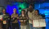 A group of five people stands in a room with camo netting, holding various props and certificates, suggesting a completed team activity or escape room challenge.