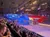 The image shows an indoor arena with stadium seating filled with people, featuring a stage area decorated with a large American flag backdrop.