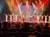 A group of performers takes a bow on stage in front of a cityscape backdrop, with audience members applauding in the foreground.