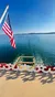 A boat with an American flag sails across a calm blue lake under a clear sky.