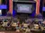 The image depicts an audience seated at tables, facing a stage with 