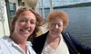 Two people are smiling and taking a selfie on a boat with a river and forested hills in the background.