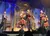 A group of musicians and singers perform energetically on stage in vintage-style outfits.
