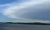 A serene view from a boat showing a wide expanse of water, distant green hills, and a partly cloudy blue sky.