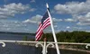 An American flag waves on a pole by a decorative railing, overlooking a body of water and a tree-lined shore under a blue sky with scattered clouds.