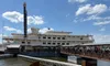 A large paddlewheel riverboat is docked beside a ramp on a sunny day, with people boarding or disembarking.