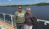 A couple is smiling and posing on a deck overlooking a large body of water, with a landscape in the background under a partly cloudy sky.