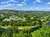 Aerial view of a lush green landscape with a river, houses, and a distant horizon under a blue sky with scattered clouds.