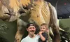 A man holding a child poses happily in front of a large dinosaur statue at a museum exhibit.