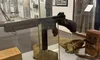 The image shows a museum exhibit displaying a World War II-era firearm, knife, and other military artifacts with informational displays and uniforms in the background.