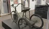 The image shows a vintage tricycle with a cargo basket on display in a museum, surrounded by historical military uniforms and memorabilia.