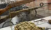 The image shows a museum display featuring a historical rifle, various military gear, and a sign labeled 