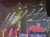 Four performers in red and black jackets are singing on stage with a vibrant light display and a graphical backdrop in front of an audience.