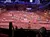 The image shows a lively Wild West-themed stage performance with dancers in period costumes and a covered wagon backdrop, set in a sandy arena with a large audience watching.